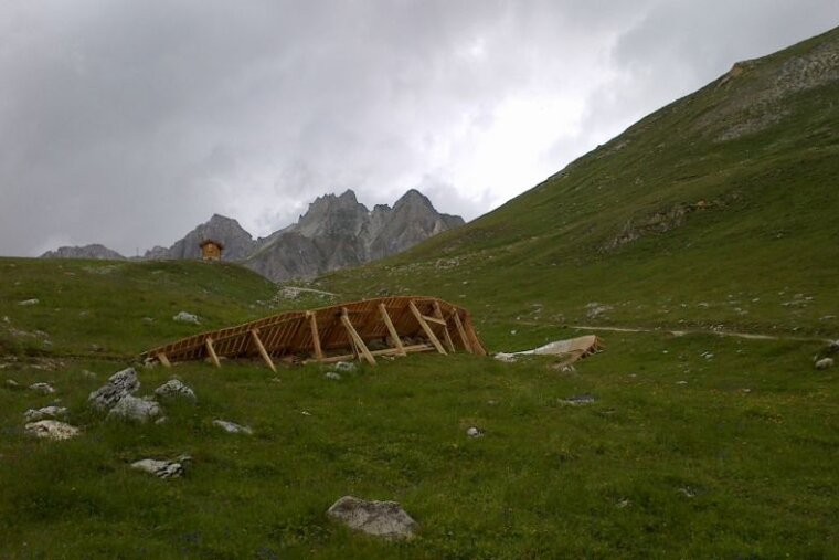 A wooden structure in the middle of a grassy field with mountains in the background