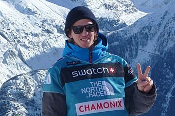 A man wearing a swatch north face shirt stands in front of snow covered mountains