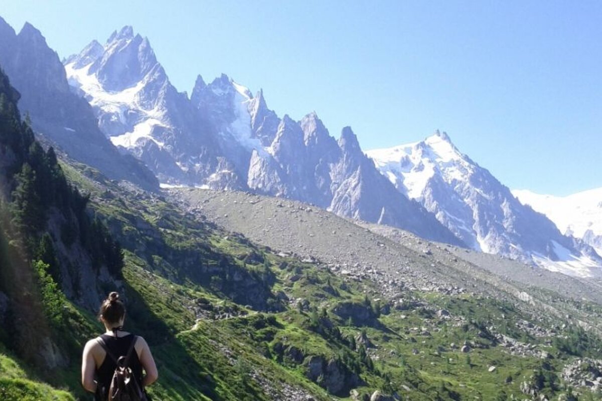 image of a woman walking along a mountain trail with the glacier on the mountain in the background, surrounded by greenery and rocks 