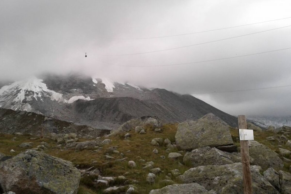 image of a cloudy sky and a cable car poking out of the cloud, the mountain with rocks and snow on and a hiking sign