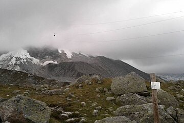 image of a cloudy sky and a cable car poking out of the cloud, the mountain with rocks and snow on and a hiking sign