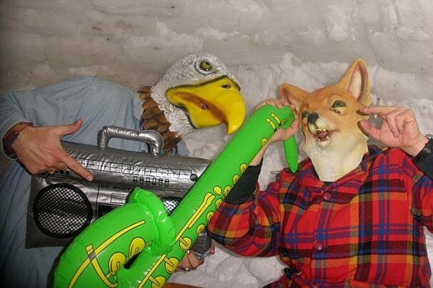 A man in a plaid shirt holds a boombox while a man in an eagle mask holds an inflatable guitar