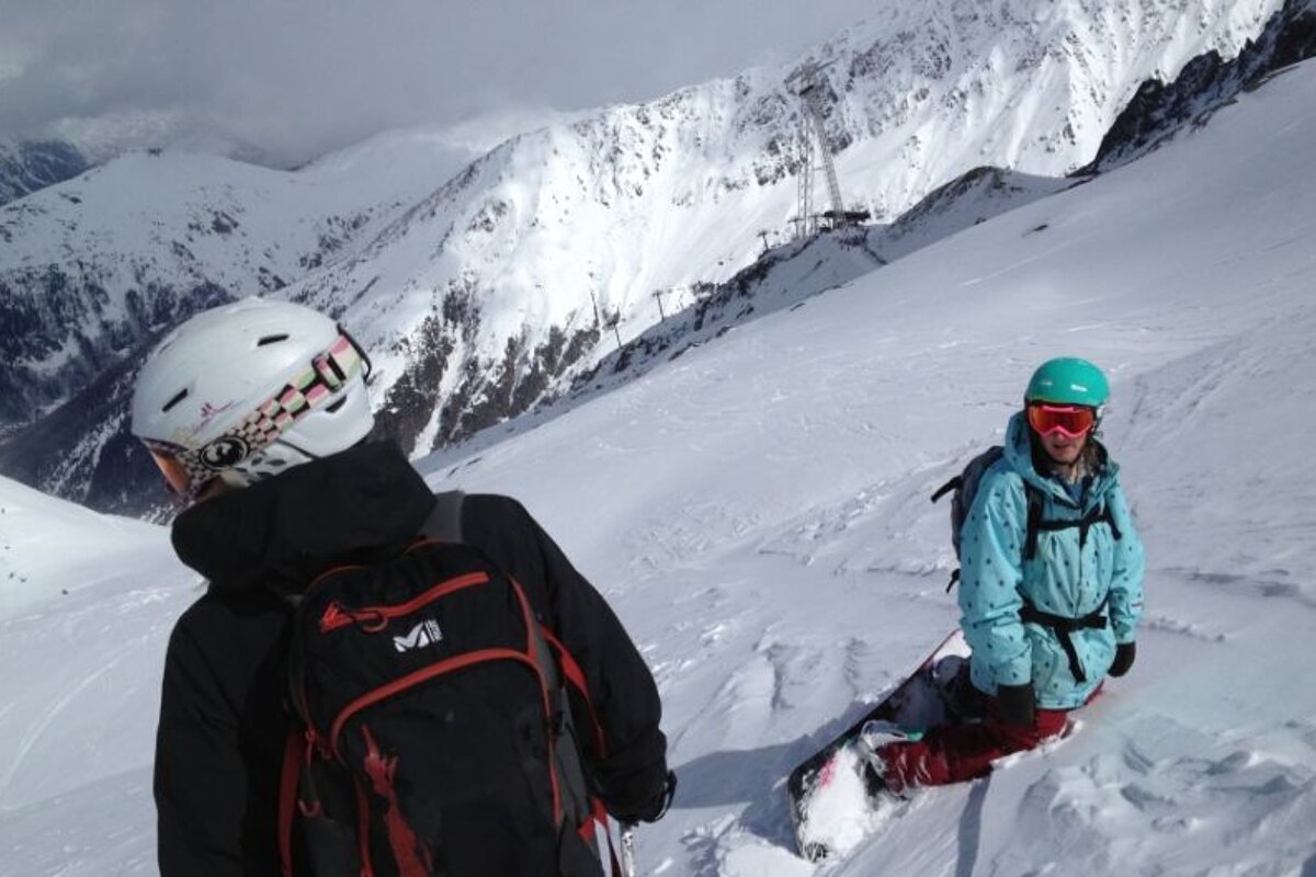 image of a snowboarder sat on the slope with a skier in front and mountains and cloud in the distance