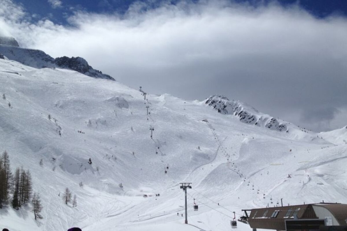 image of a piste with some lifts and runs and people skiing down them with blue sky and a few clouds