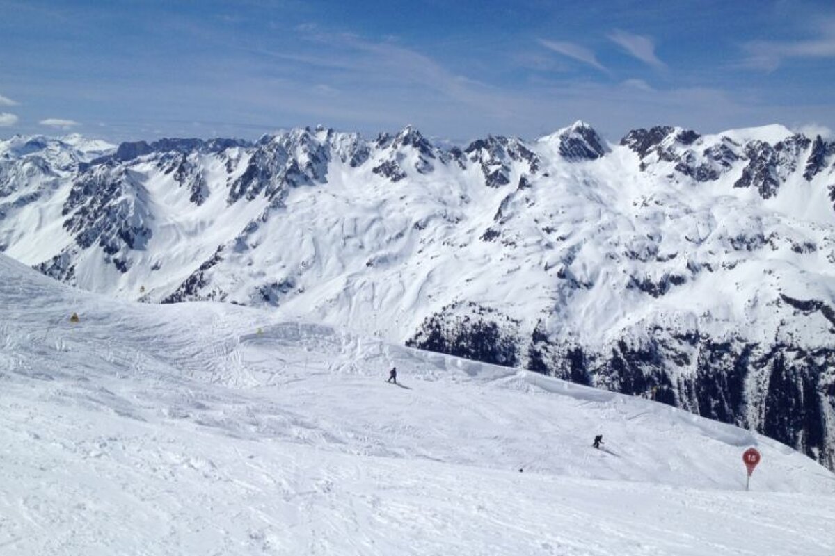 image a piste with skiiers on and snowy mountains in the distance with blue skies