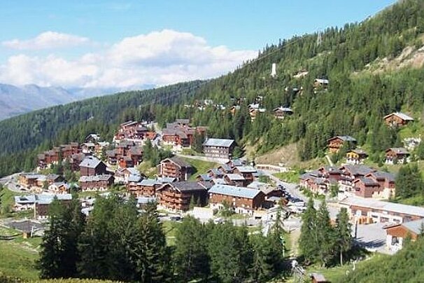 image of a town up a mountain surrounded by green trees and mountains in the distanc, with blue sky and some clouds above