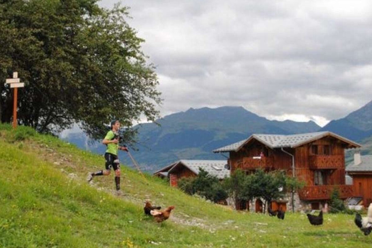 image of a man hiking downhill with some chalets and mountains in the background and chickens