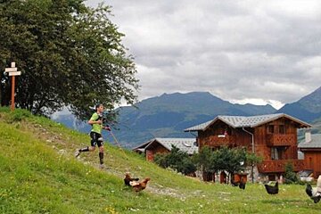 image of a man hiking downhill with some chalets and mountains in the background and chickens
