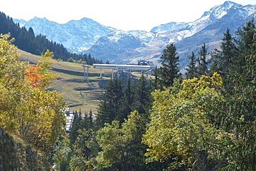 image of a chairlift tucked behind trees in the summer with the mountains behind and snow on the mountain peaks