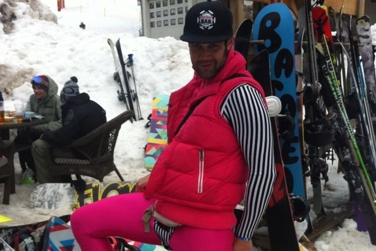 A man wearing a pink vest and a black hat stands in front of a snowboard with the letter b on it