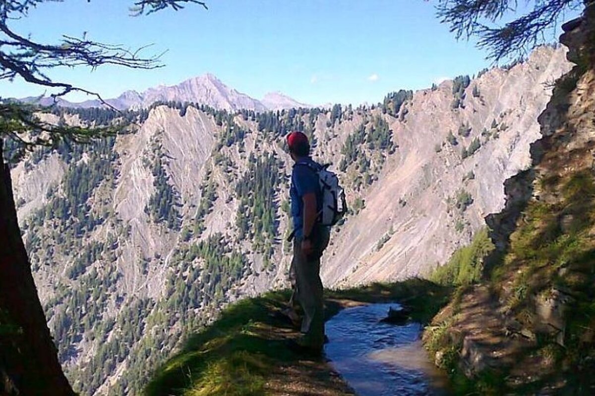 image of a man stood on a footpath on a mountain side in the shade, looking at the rocky and tree covered mountains in the distance