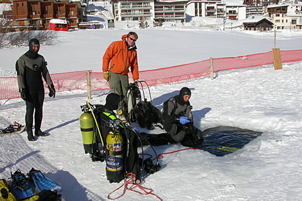 Three scuba divers are getting ready to dive into a hole in the snow
