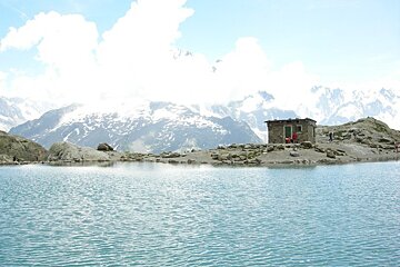 A small house sits on the shore of a lake with mountains in the background