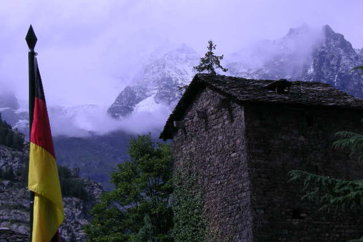 A german flag is flying in front of a stone building with mountains in the background