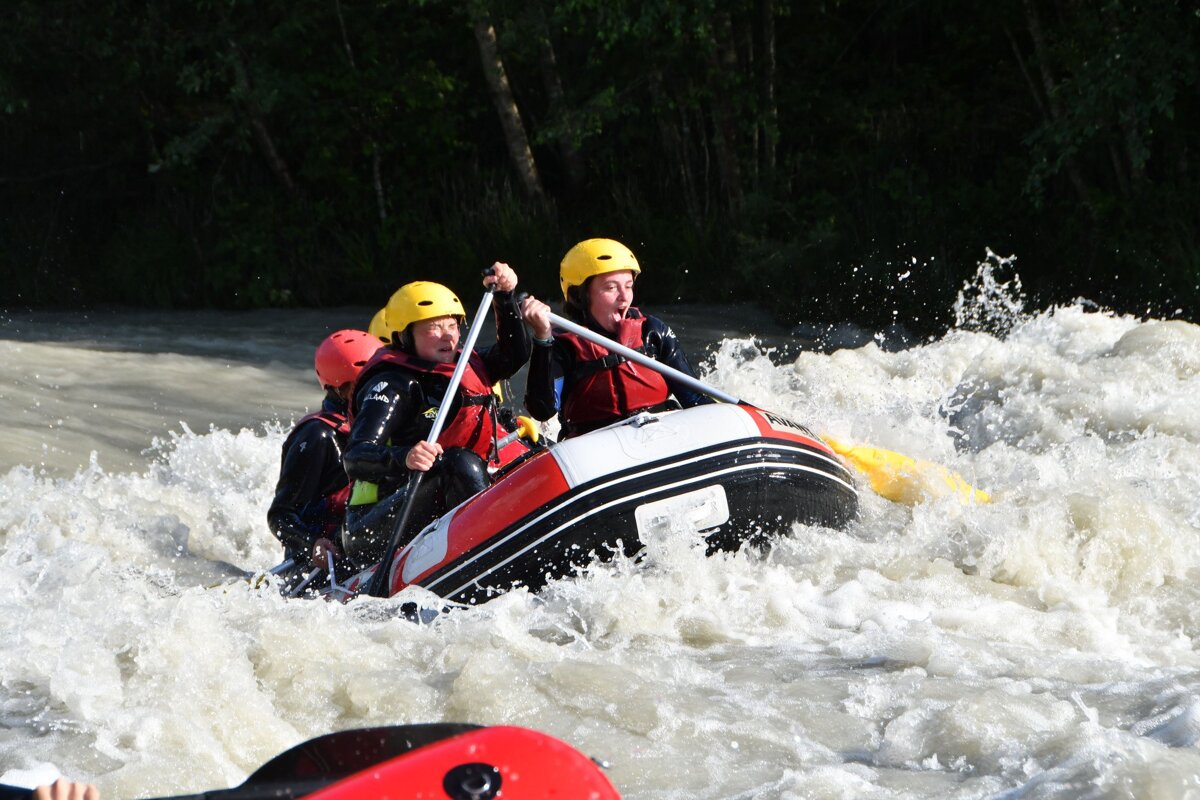 Rafters in helmets and life vests navigate a red and black raft through turbulent white water, paddling hard amidst splashes.
