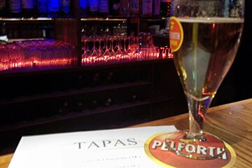 A glass of beer sits on a table next to a menu for tapas