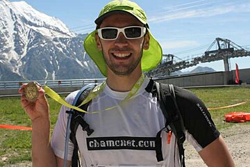 A man wearing sunglasses and a green hat is holding a medal in front of a mountain
