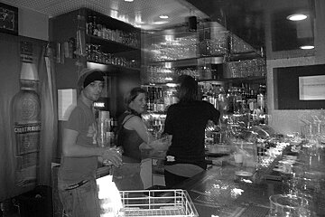 A man and a woman standing in a bar with a bottle of chaterousse behind them
