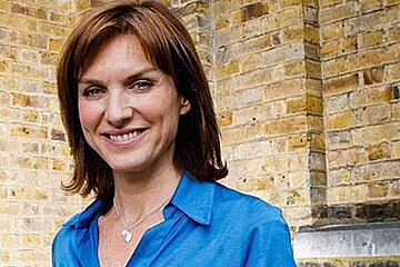 A woman in a blue shirt and necklace is smiling in front of a brick wall