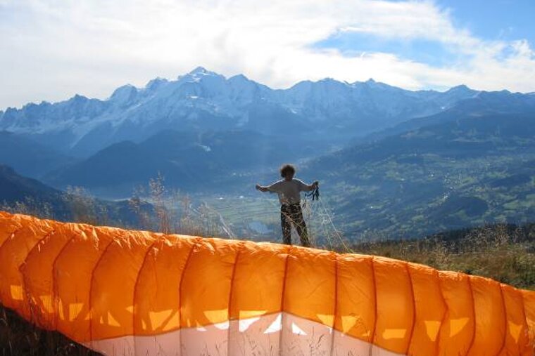 A man is standing next to an orange parachute with mountains in the background