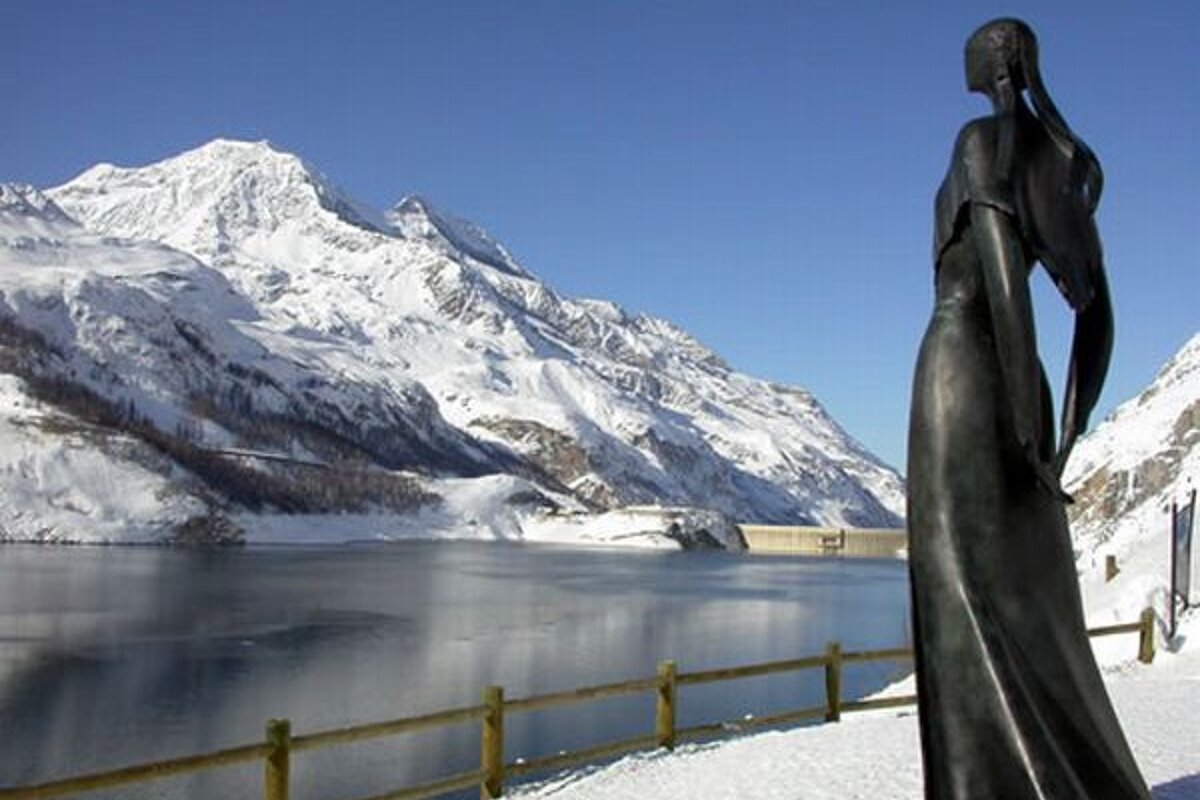 A statue of a woman stands in front of a snowy mountain and lake