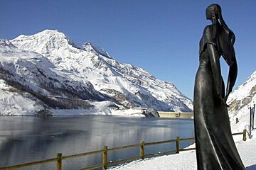 A statue of a woman stands in front of a snowy mountain and lake