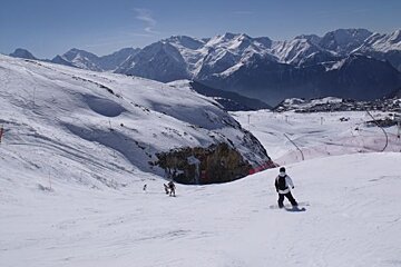 A snowboarder rides down a snowy slope with mountains in the background