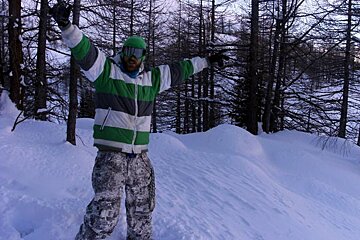 A man in a green and white striped jacket stands in the snow with his arms outstretched