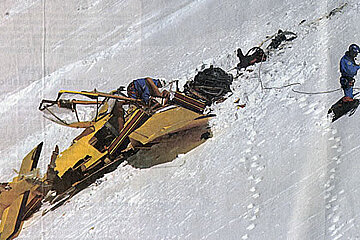 A snowmobile is being pulled up a snowy hill by a man on skis