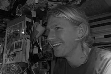 A black and white photo of a smiling man in front of a wall with a calendar that says ' sydney ' on it