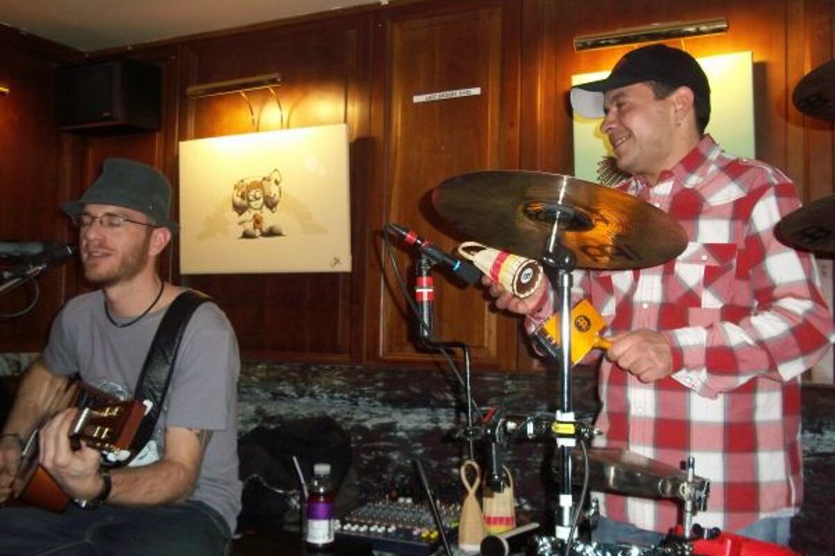 Two musicians perform live indoors. A guitarist sings, while another plays percussion (shakers, cymbals) in a red plaid shirt.