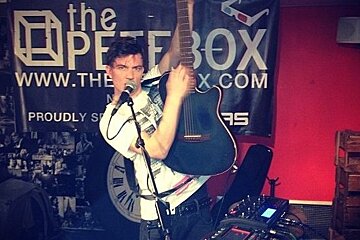 A man singing into a microphone in front of a sign that says the pete box