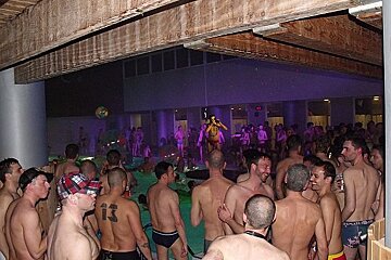 A crowded indoor pool party with many shirtless men. Dim purple lighting, wooden beams overhead, and a person on a platform in the background.