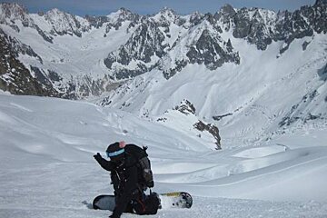 A person is kneeling down in the snow with a snowboard in front of a snowy mountain