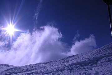 The sun shines brightly through the clouds above a snowy slope