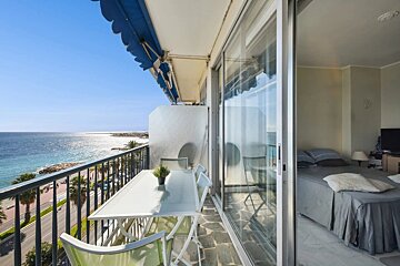 Balcony with dining set and blue awning, overlooking a sunny ocean, coastal road, and palm trees. Sliding door opens to a bedroom.