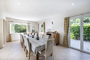 Bright dining room featuring a long table with upholstered chairs, yellow curtains, and glass doors opening to a green garden patio.