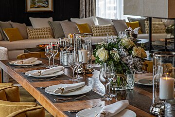 An elegant dining table is set with white plates, glasses, candles, and a floral centerpiece, facing a cozy living room with a sofa and cushions.
