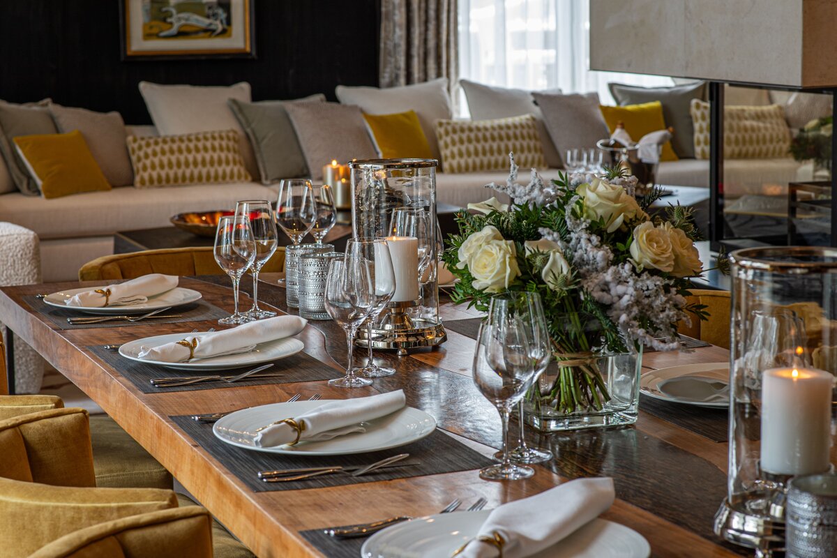 An elegantly set wooden dining table with candles and white roses, looking toward a cozy, modern living room with a plush sofa.