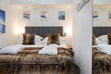 Cozy bedroom with a grey headboard, faux-fur pillows and blanket, and mountain art prints on the white wall.