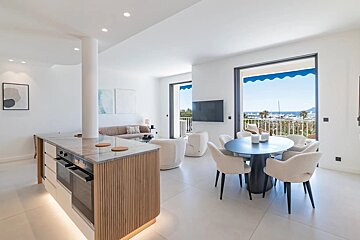 Bright, modern open-plan interior with a kitchen island, dining set, living area, and balcony overlooking a scenic marina.