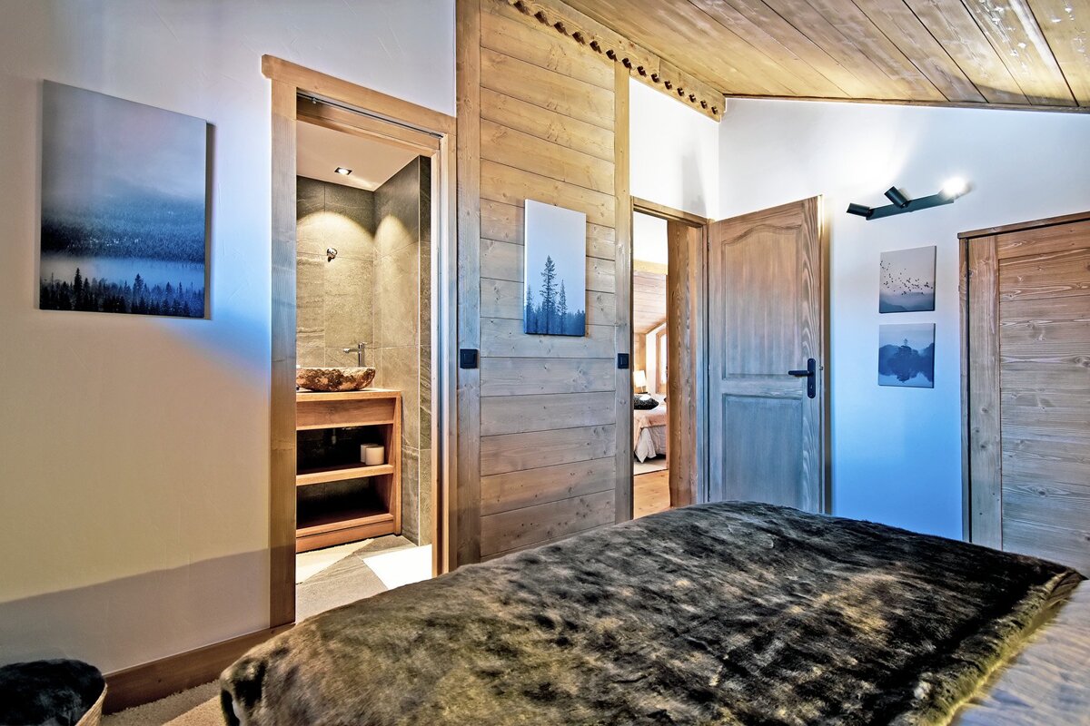 A cozy cabin-style bedroom featuring wood paneling, a fur-textured blanket, nature wall art, and a view into an en-suite bathroom.