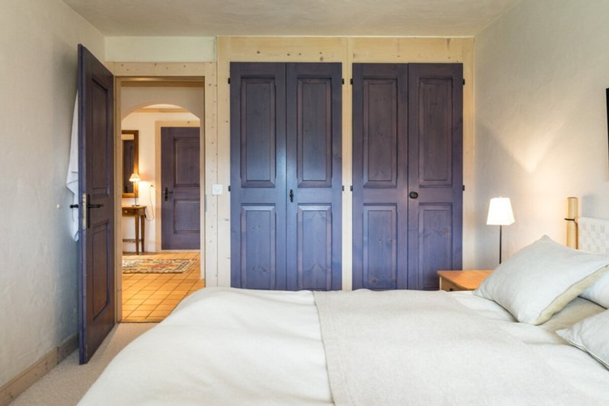 A cozy bedroom featuring a white-dressed bed, purple wooden closets, and an open door looking into a warm hallway.
