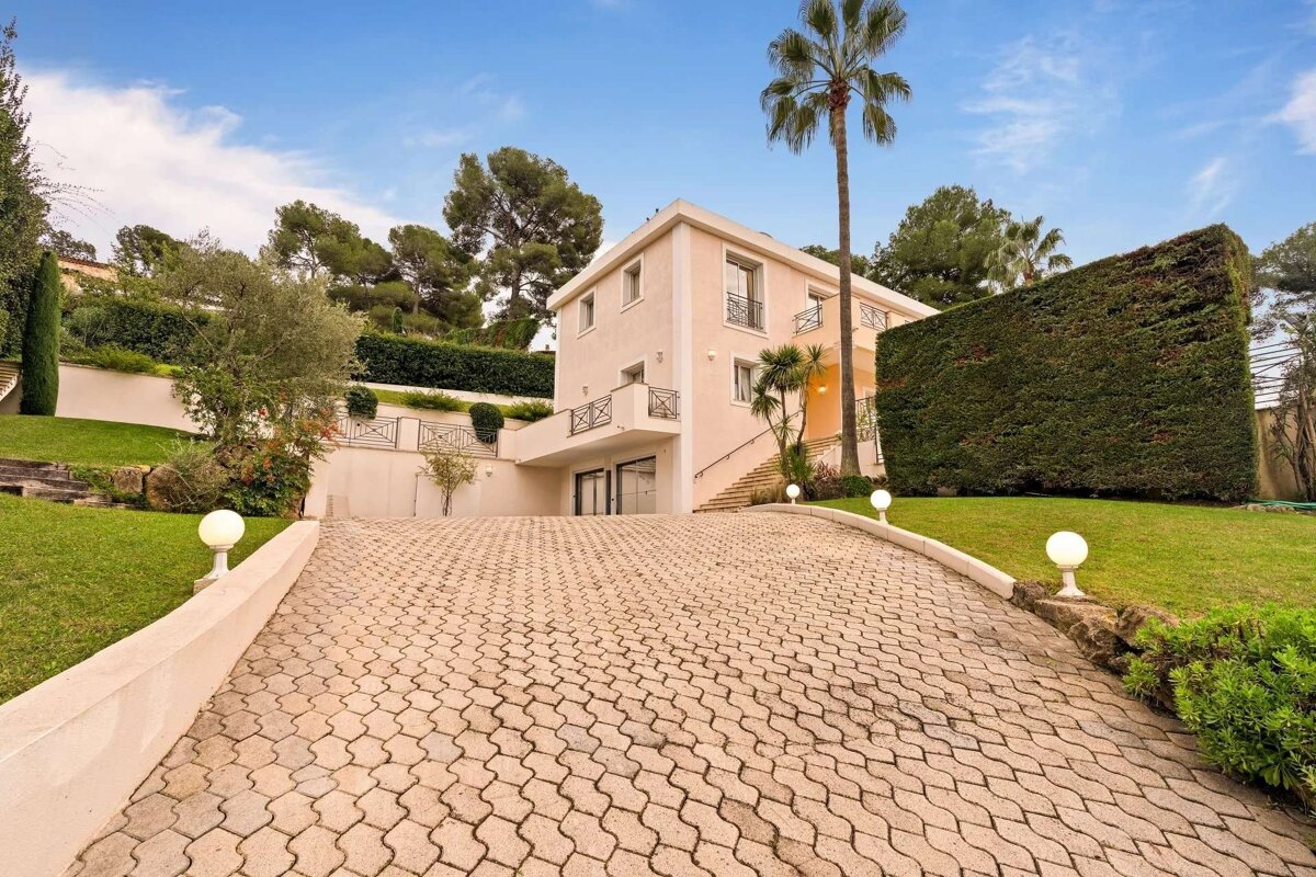 A grand, light-colored house with a long, patterned paved driveway, lush green gardens, hedges, and tall trees, including a prominent palm, under a blue sky.
