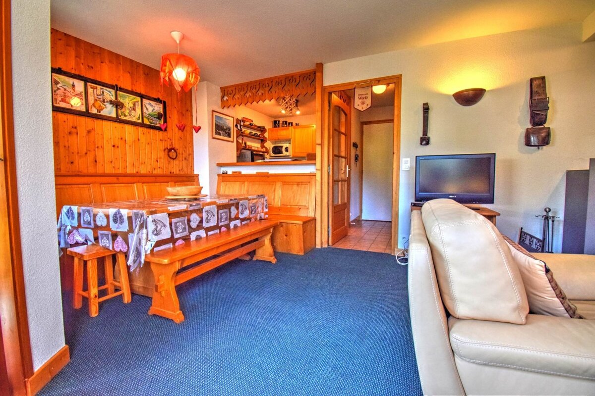 A warm, chalet-style living space featuring a wooden dining nook, blue carpet, kitchenette, and a cream sofa with a TV.