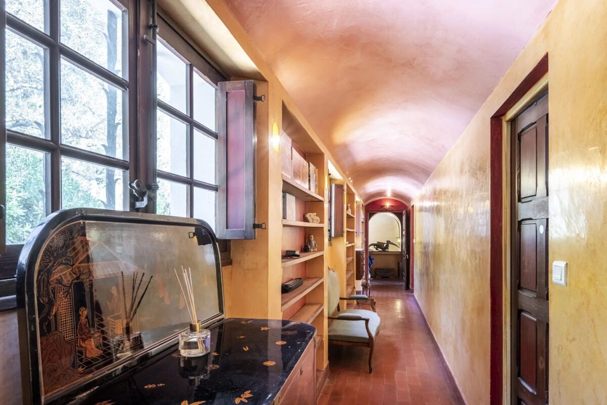 A narrow corridor with a pink vaulted ceiling, warm yellow walls, windows, bookshelves, and an ornate dark cabinet in the foreground.