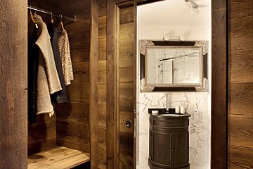 A rustic wooden wardrobe with clothes on the left, opens to a luxurious bathroom on the right, featuring a dark wood vanity, ornate mirror, and white marble.