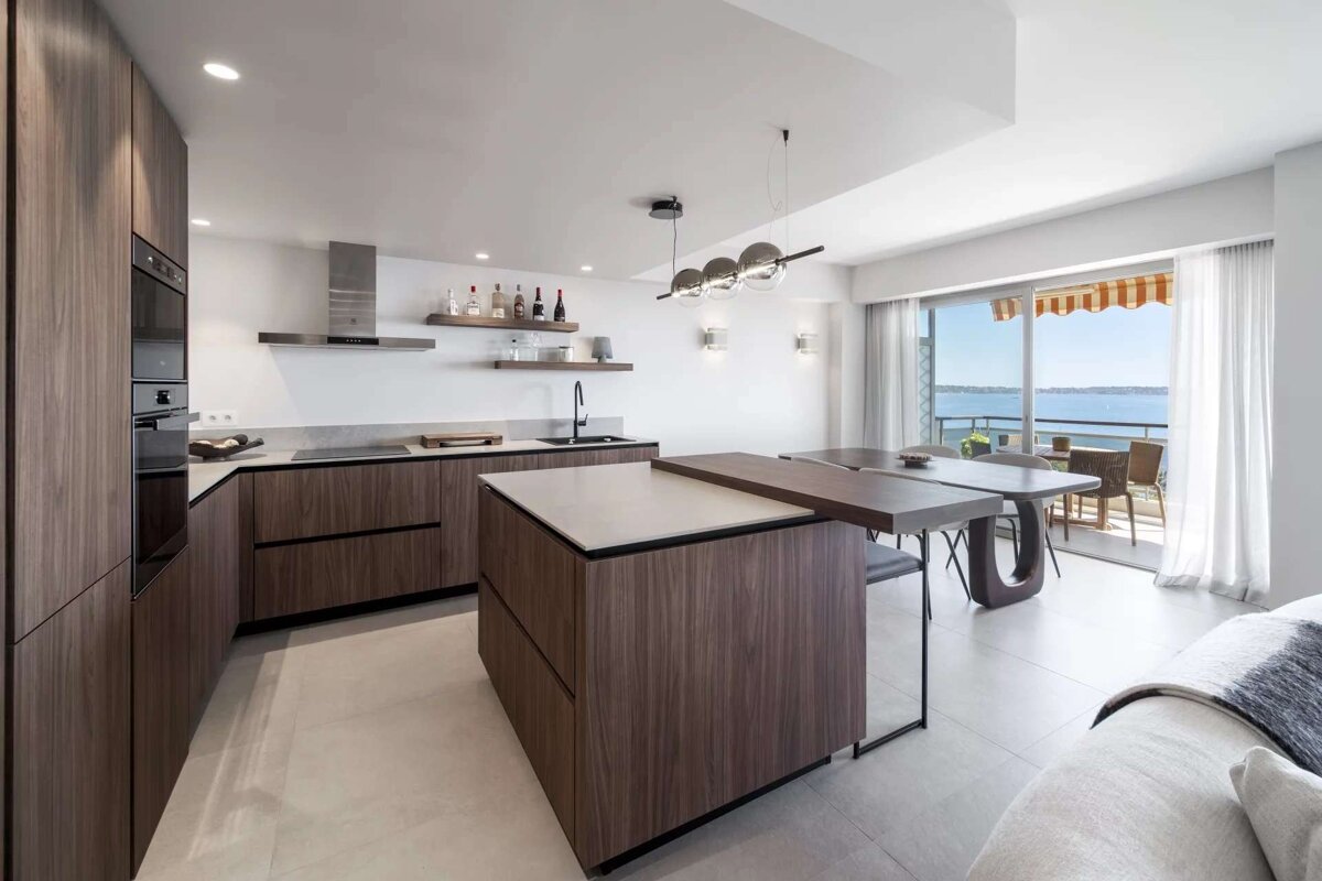 A sleek, modern kitchen with dark wood cabinets and island features an integrated dining table and a balcony overlooking a scenic sea view.