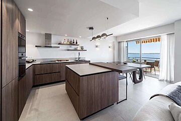 A sleek, modern kitchen with dark wood cabinets and island features an integrated dining table and a balcony overlooking a scenic sea view.