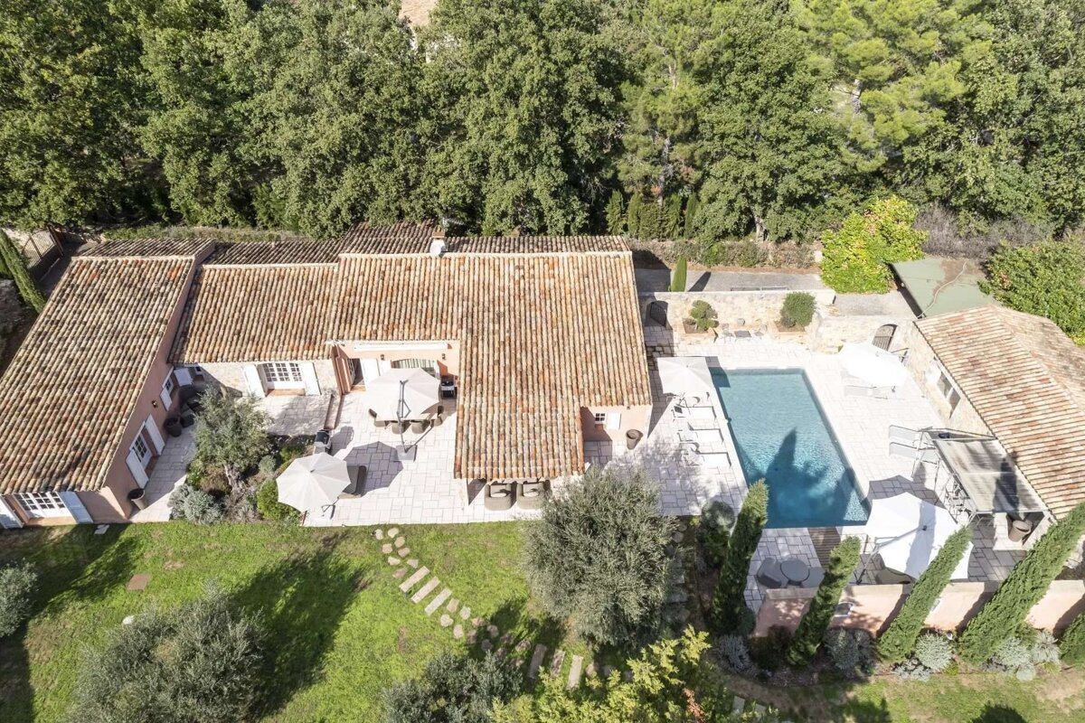 Drone shot of a Mediterranean-style home with red tile roofs, a clear blue pool, paved patios, and a verdant garden, seen from above.